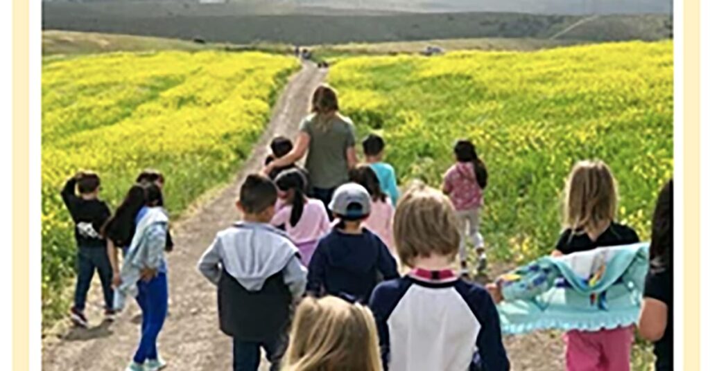 A group of children and a woman stroll down a dirt path lined with vibrant yellow flowers and green hills under a cloudy sky, reminiscent of an idyllic Mailchimp campaign banner. The children, dressed in casual attire, clutch jackets or blankets as they explore the scenic route.