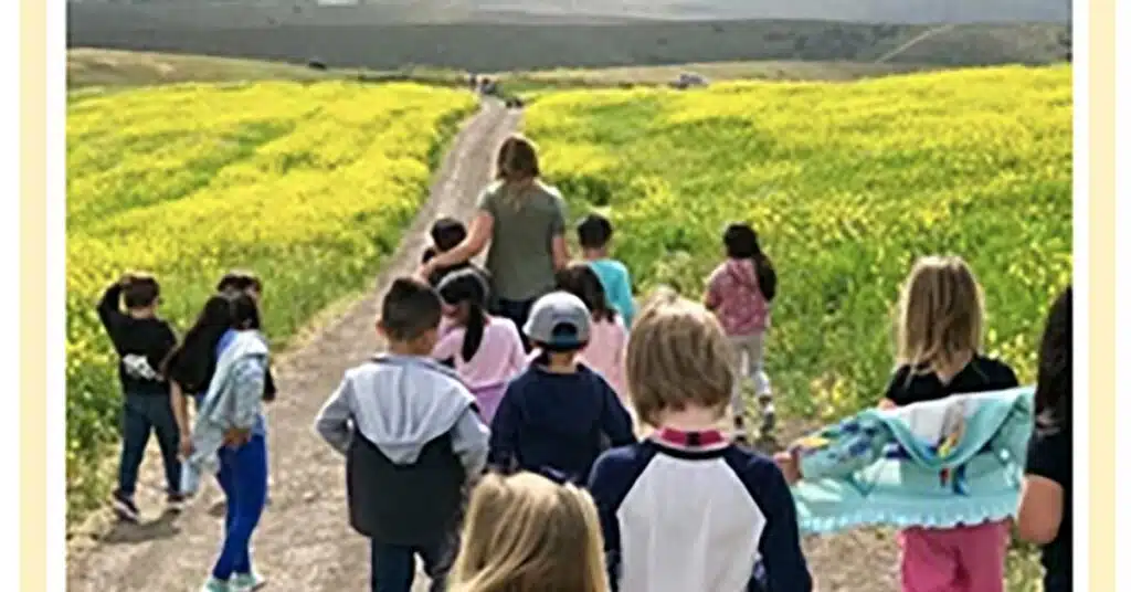 A group of children and a woman stroll down a dirt path lined with vibrant yellow flowers and green hills under a cloudy sky, reminiscent of an idyllic Mailchimp campaign banner. The children, dressed in casual attire, clutch jackets or blankets as they explore the scenic route.