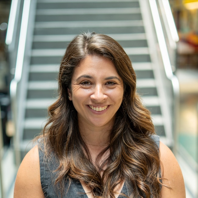Michelle Jaconette, a woman with long brown hair and a gray top, smiles at the camera while standing in front of a blurred staircase indoors.