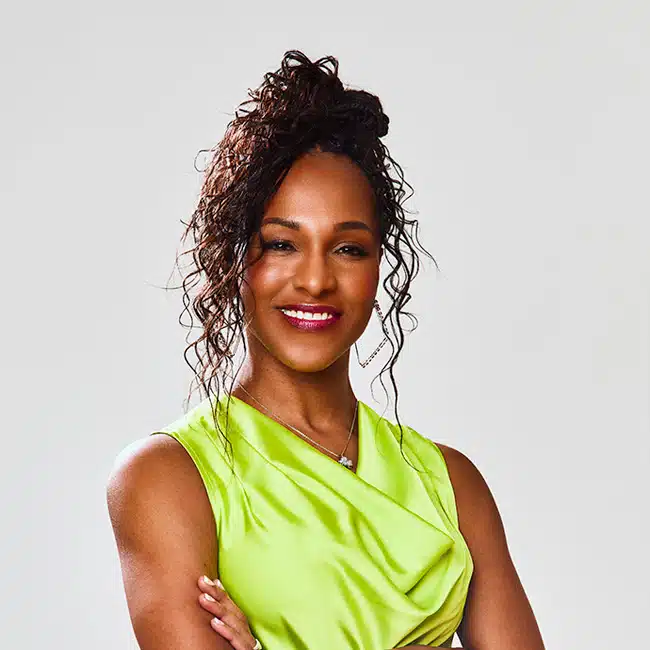 Michelle Pledger, a woman with curly hair styled in an updo, wears a sleeveless lime green top and smiles confidently with arms crossed in front of a plain light background.