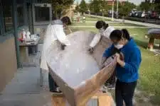 Three masked individuals collaborate on a large fiberglass structure outdoors. A person in a blue hoodie stands to the right as two others, clad in lab coats, focus on the interior. Supplies are neatly arranged on a nearby table, ready for action.