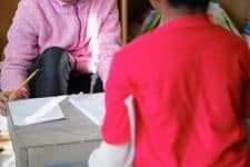 Two children are seated and working with pencils and paper. One child wears a pink sweater and holds a pencil, while the other wears a red shirt. Papers and a box are visible between them.