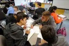 A group of students is sitting around a table, collaborating and discussing like theyre curating the perfect SEO keywords for a project. Surrounded by papers, notebooks, and a smartphone, the busy classroom buzzes with other students diligently working in the background.