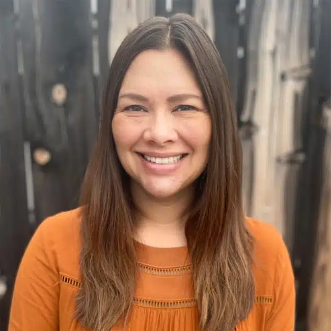 A person with long brown hair is smiling, standing by their point of contact—a rustic wooden fence. They are wearing a vibrant orange top.