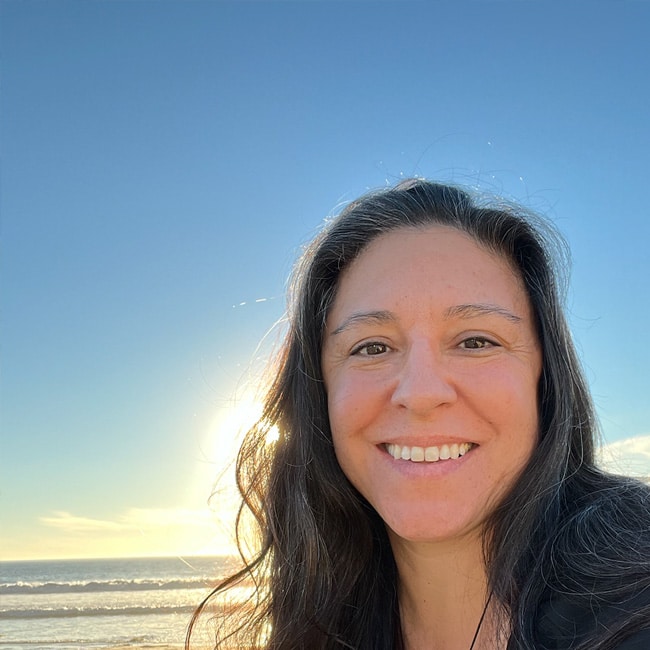 Nikki Hinostro, with long dark hair, smiles at the camera in front of a beach, the sun shining brightly in the clear blue sky behind her.