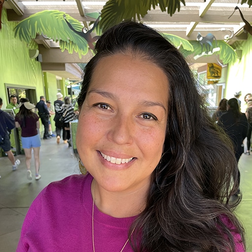 Nuvia Ruland, a woman with long dark hair, smiles at the camera while wearing a purple top. She is indoors with people walking around, greenery, and animal decorations in the background. Bright, natural light fills the space.