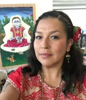 A person with long dark hair, wearing a red patterned top and earrings, stands linked to vibrant artwork featuring a skeleton in traditional clothing. A red flower adorns their hair.