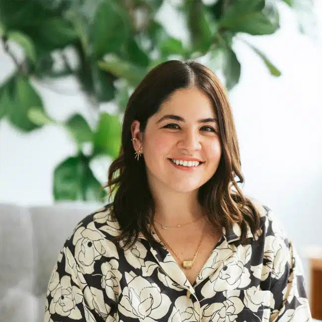 Paola Capo-Garcia, a woman with shoulder-length brown hair, smiles while sitting in front of green leafy plants. She is wearing a floral patterned shirt and gold jewelry that perfectly complements her radiant demeanor.