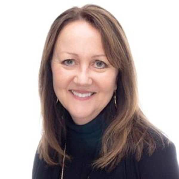 A woman with long brown hair, smiling, is wearing a black top and standing against a plain white background.