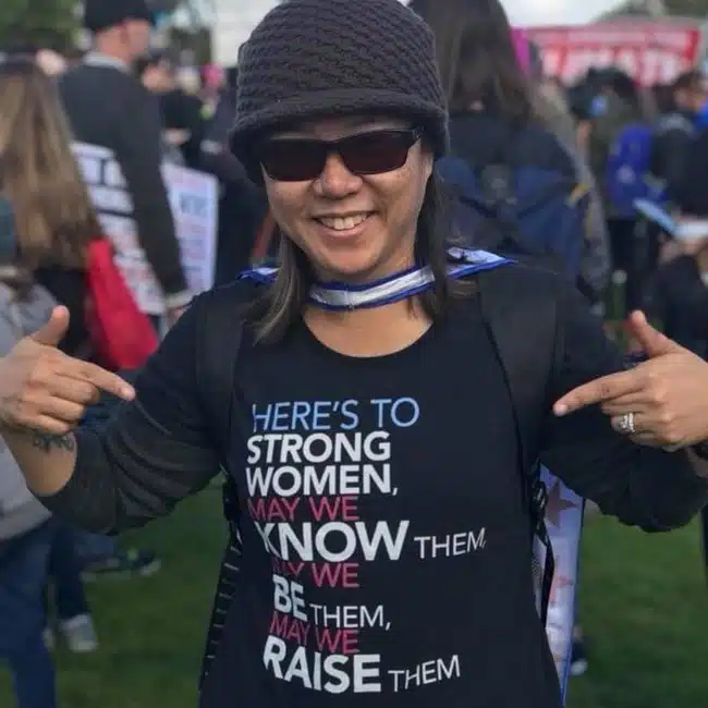 A person smiles and points to a shirt with the text: Heres to strong women, may we know them, may we be them, may we raise them. They are wearing sunglasses and a knitted cap in a crowd of people.