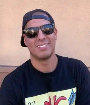 A person wearing a backwards baseball cap, sunglasses, and a dark shirt with a colorful graphic smiles while sitting outdoors against a beige background.
