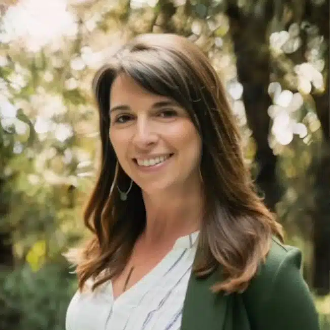 Stacey Lopaz, a woman with long brown hair, smiles in a lush green outdoor setting. She is wearing a white blouse and a green jacket as sunlight filters through the trees in the background.