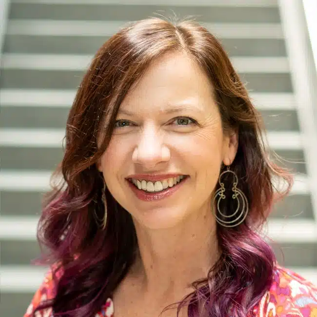 A woman with long, wavy brown hair and purple highlights smiles at the camera. She is wearing large hoop earrings and a colorful top, standing in front of a blurred stairway background.