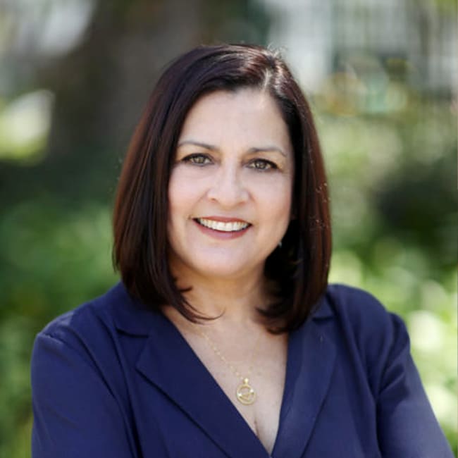 A person with shoulder-length dark hair smiles at the camera. They are wearing a navy blue blazer with a gold necklace. The background is a blurred outdoor setting with greenery.