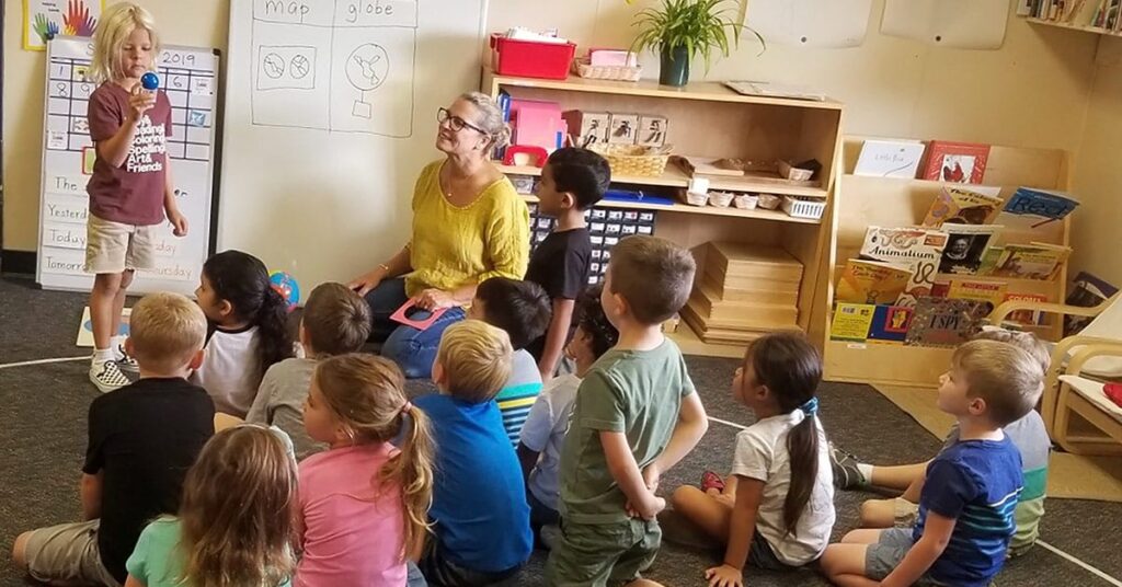A group of young children sit on the floor, attentively listening to a smiling teacher who is kneeling and guiding them. A child stands next to a calendar, speaking and pointing to the display. The colorful classroom fosters positive relations, with shelves full of books and educational materials.