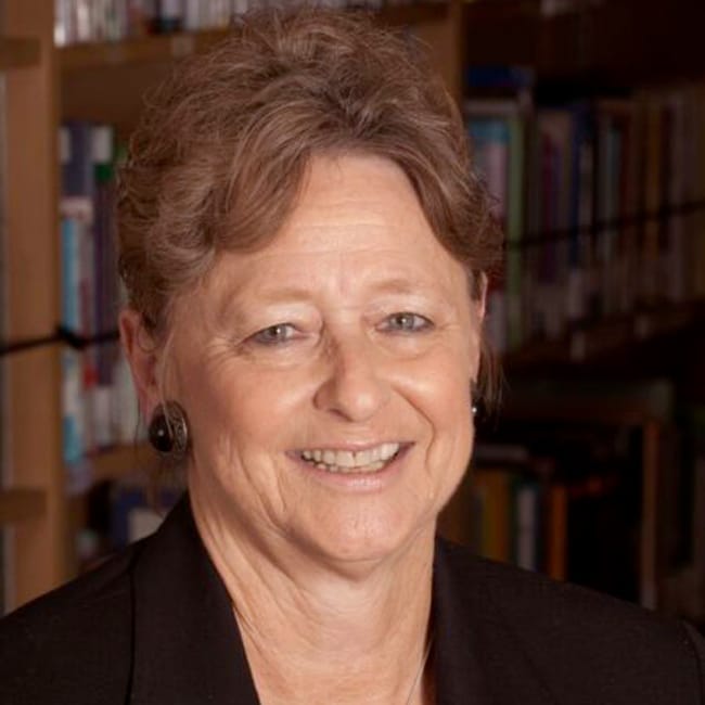 A smiling woman with short, curly hair is standing in front of a bookshelf filled with books. She is wearing a black top and earrings. The background suggests a library or study setting.