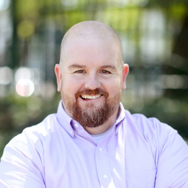 A person with a beard smiling, wearing a light purple button-up shirt, gives off a cinematic vibe as if stepping right out of a film reel. The background is blurred, featuring trees and greenery.