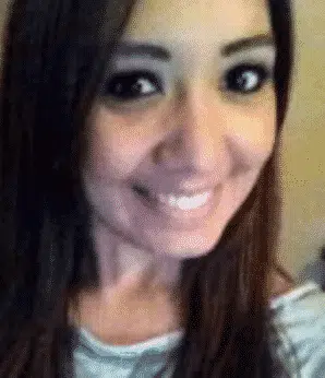 A person with long brown hair and dark eyes is smiling widely. They are wearing a light-colored top and are positioned slightly off-center, looking directly at the camera.