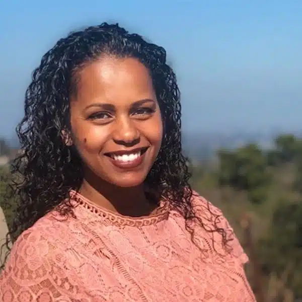 A person with curly hair smiles at the camera. They are wearing a pink lace top. The background shows a blurred outdoor setting with greenery and a blue sky.
