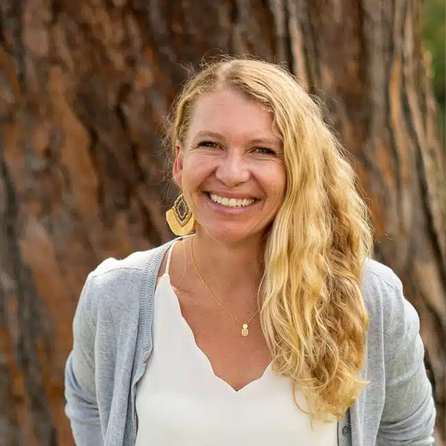 A smiling person with long, wavy blonde hair stands outdoors in front of a tree trunk. They are wearing a light gray cardigan, a white blouse, and a pendant necklace, with a geometric earring visible on one ear.