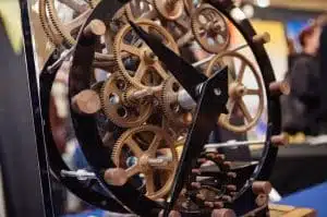 Intricate display of interconnected wooden gears and cogs on a stand, with a dark metal frame. The background is blurred, hinting at an indoor setting with colorful elements.