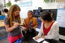 In a lively classroom scene, three students collaborate on a project. One student holds a tool while another leans in to observe and the third diligently takes notes. They are surrounded by educational materials and equipment, creating an immersive learning environment focused on problem-solving issues.