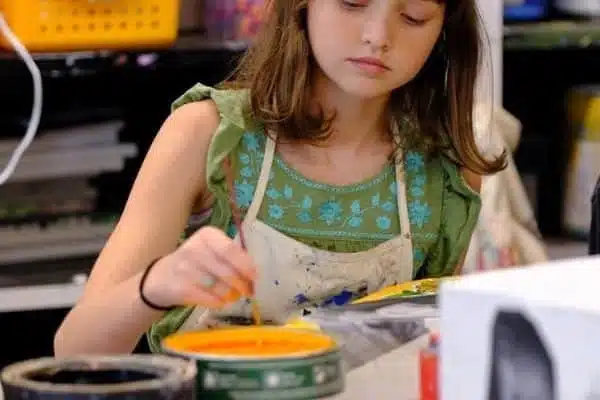 A young girl, embracing her artistic spirit, concentrates on painting in sepia tones. Wearing a green top and an apron, she sits at a table with art supplies, mixing colors with her brush.