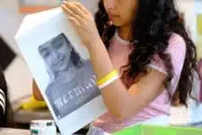 A person with long hair wearing a pink shirt is holding up a grayscale printed image of a face. A yellow wristband is visible on their wrist. The background is blurred, with indistinct objects and colors.