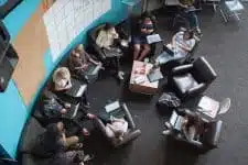 Overhead view of a group of people sitting in a circle on armchairs and sofas, engaged in a discussion or meeting. They are using laptops and notebooks, with a large whiteboard visible on the wall. The room has a casual and collaborative atmosphere.