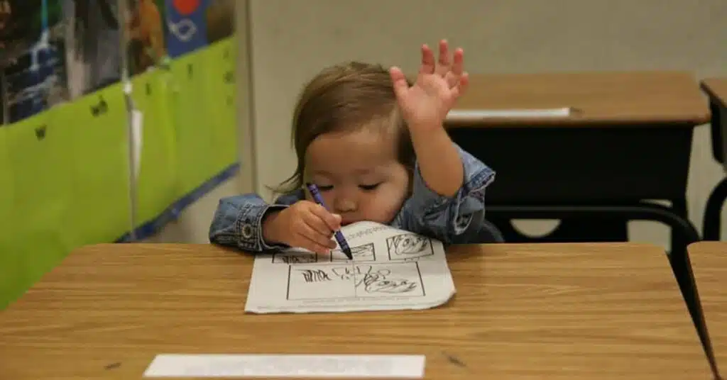 A young child with long hair sits at a desk, holding a pen and writing on a paper, reminiscent of capturing thoughts for a podcast collection. With one hand raised, wearing a denim jacket, the classroom walls provide the backdrop to this moment of creativity.