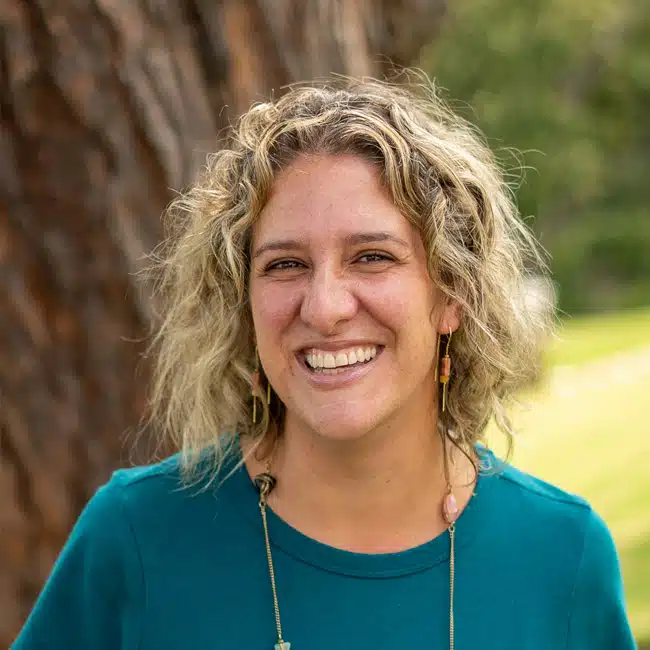 Tina Schuster, with her curly hair and a radiant smile, stands outdoors in a teal top and earrings. She poses gracefully in front of a tree, surrounded by lush greenery and a beautifully blurred landscape that hints at the serenity of nature.