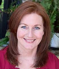 A woman with shoulder-length red hair is smiling at the camera, exuding a friendly vibe. She is wearing a maroon top and seated in front of a lush background with green plants, reminiscent of an inviting Spotify playlist cover.
