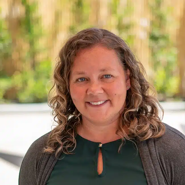 A person with curly hair wearing a green blouse and gray cardigan smiles outdoors, with a blurred natural background, perhaps sharing info for their latest podcast episode.