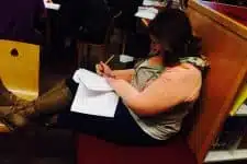 A person is sitting on a cushioned bench in a library, writing on papers with a pencil. They have their legs resting on a nearby chair. Shelves filled with books can be seen in the background.