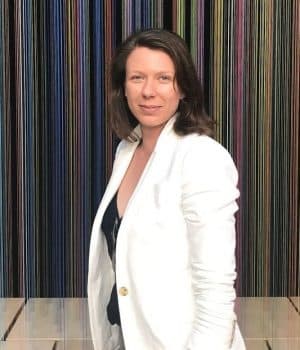 A person with shoulder-length brown hair wearing a white jacket stands in front of a vibrant global-inspired striped background, seamlessly blending into the scene like a dynamic webview capturing diverse cultures.