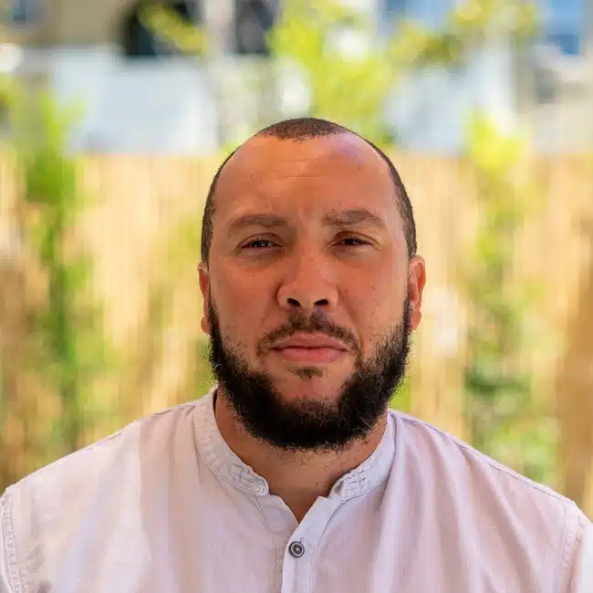 A man with a beard and a shaved head is wearing a white shirt. He is outdoors with greenery and a wooden fence in the background. The lighting is bright and natural.