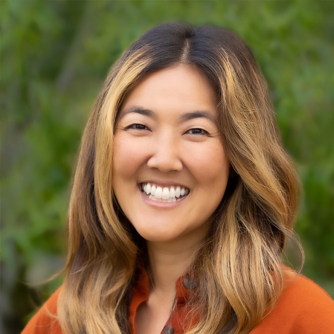 A woman with long, wavy brown hair and blonde highlights smiles outdoors, wearing an orange shirt. The background is blurred greenery.