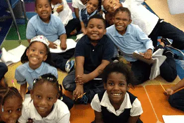 A group of cheerful schoolchildren in blue and white uniforms sit together on a colorful classroom floor, smiling for the camera. They appear engaged and happy, like an episode of youthful joy unfolding amidst notebooks and papers around them.