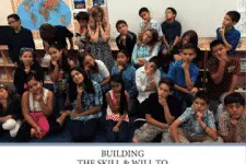 A group of children in a classroom posing for a photo, some sitting and some standing, all with thoughtful expressions. A world map is visible on the wall behind them. Text at the bottom reads Building the Skill & Will To...