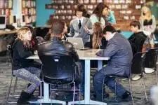 In a classroom discussion set against a dark background, students and a teacher engage dynamically. While some listen attentively and others focus on their laptops, the atmosphere hums with curiosity, almost like observing scorpions under ultraviolet light.