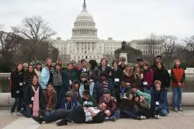 A diverse group of students poses in front of the United States Capitol building. They are wearing casual clothing and many have name tags. The sky is overcast and trees are visible in the background.