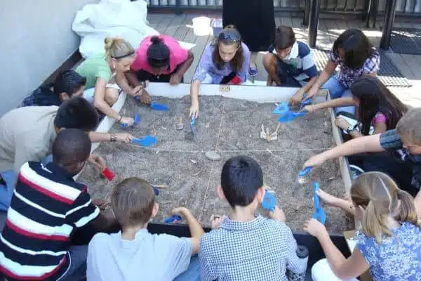 A group of children attentively engage in a simulated archaeological dig. They kneel around a sand pit, using small tools to uncover and explore items in a grid-marked area. The activity takes place outdoors on a sunny day.