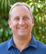 A person with short, light-colored hair is smiling broadly in front of a green, leafy background. They are wearing a blue polo shirt.