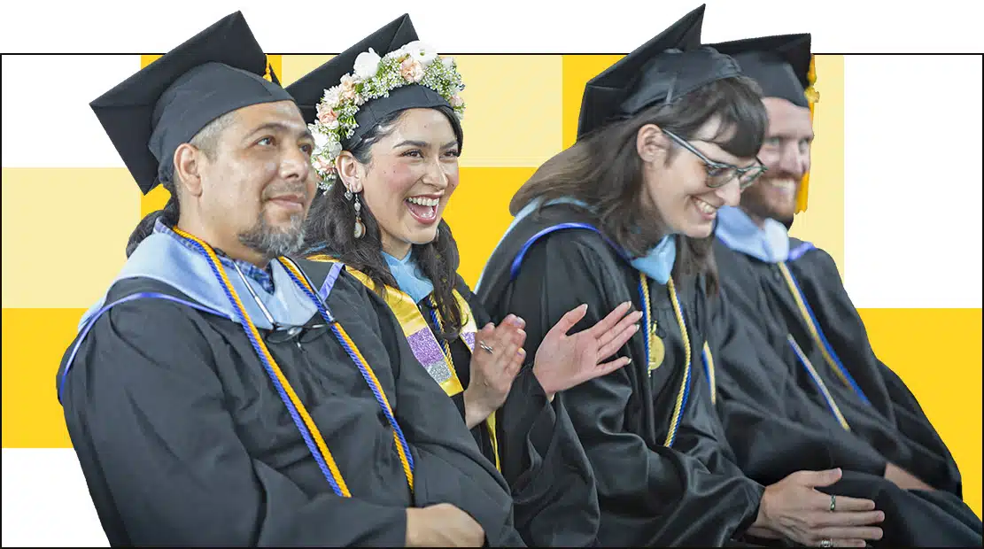 A group of graduates in caps and gowns sit in a row, smiling and clapping. One woman in the center wears a flower crown as they celebrate their achievements. A yellow and white checkered pattern serves as the backdrop, highlighting the vibrant programs theyve completed.