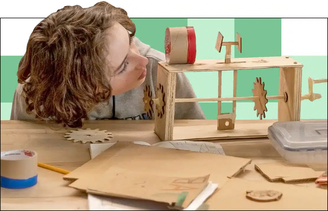 A child with curly hair is observing a wooden mechanical structure with gears on a table covered in papers, cardboard, and tape. The background has a green and white checkered pattern.