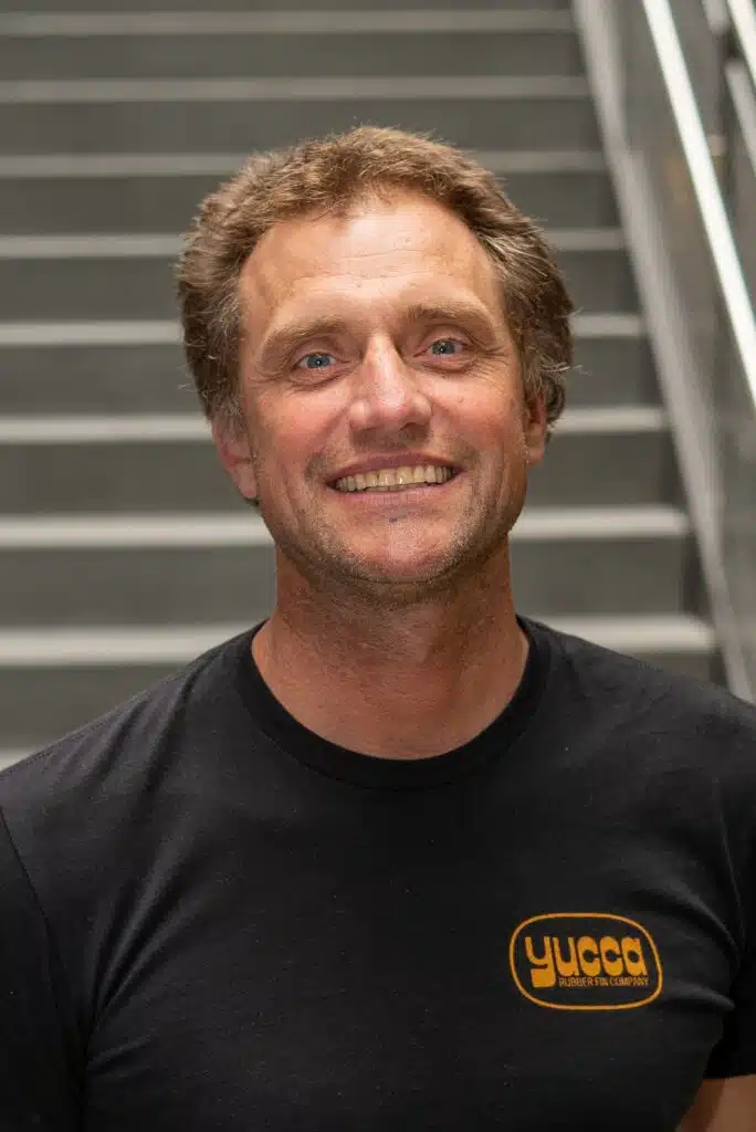 A man with short, curly hair smiles at the camera, standing indoors in front of a staircase. He is wearing a black T-shirt with the orange logo yucca.