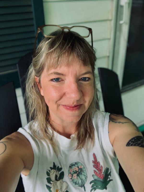 A person with shoulder-length blonde hair and tattoos on their arms is smiling at the camera. They are wearing a sleeveless white shirt adorned with plant doodles and have sunglasses resting on their head. The scene, captured in a sepia tone, shows them sitting by a green and white wall.