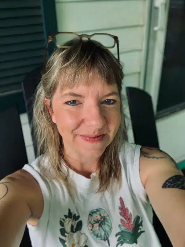 A person with shoulder-length blonde hair and tattoos on their arms is smiling at the camera. They are wearing a sleeveless white shirt adorned with plant doodles and have sunglasses resting on their head. The scene, captured in a sepia tone, shows them sitting by a green and white wall.