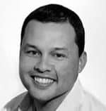 Portrait of a smiling person with short dark hair, wearing a light-colored shirt. The image is in black and white.