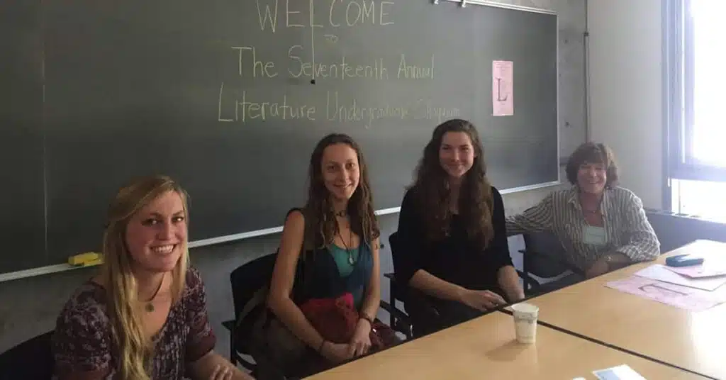 Four people sit at a table in a classroom with a chalkboard behind them. The board reads, Welcome to the Seventeenth Annual Literature Undergraduate Colloquium. The group appears to be participating in or preparing for an academic event.